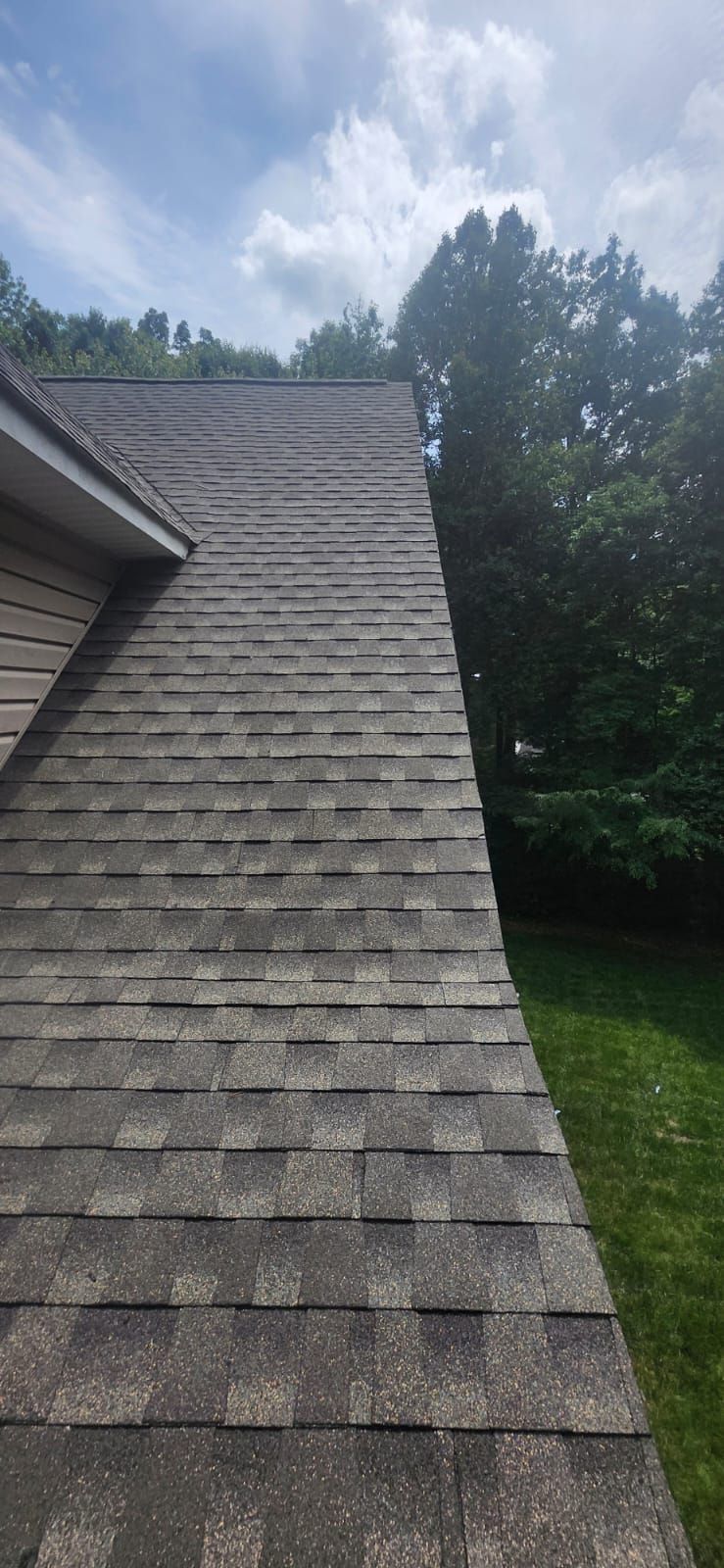 A roof covered with gray asphalt shingles against a cloudy sky and green trees.