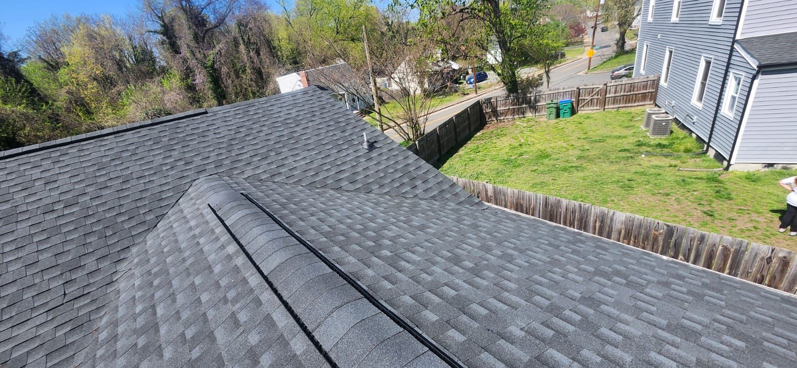View from a dark grey shingled roof, looking towards green grass and a wooden fence, with blue sky.