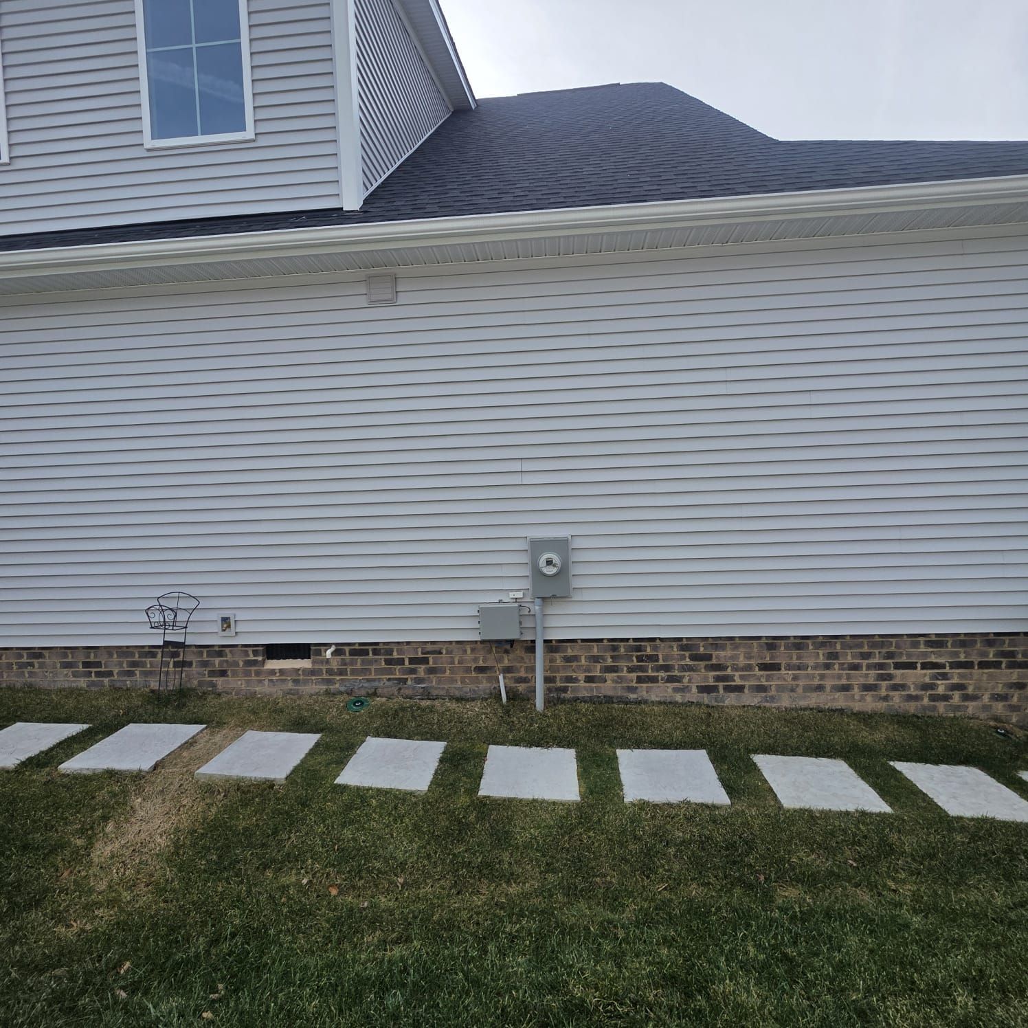 Side of a house with gray siding, brick foundation, and a row of square stepping stones in the grass.