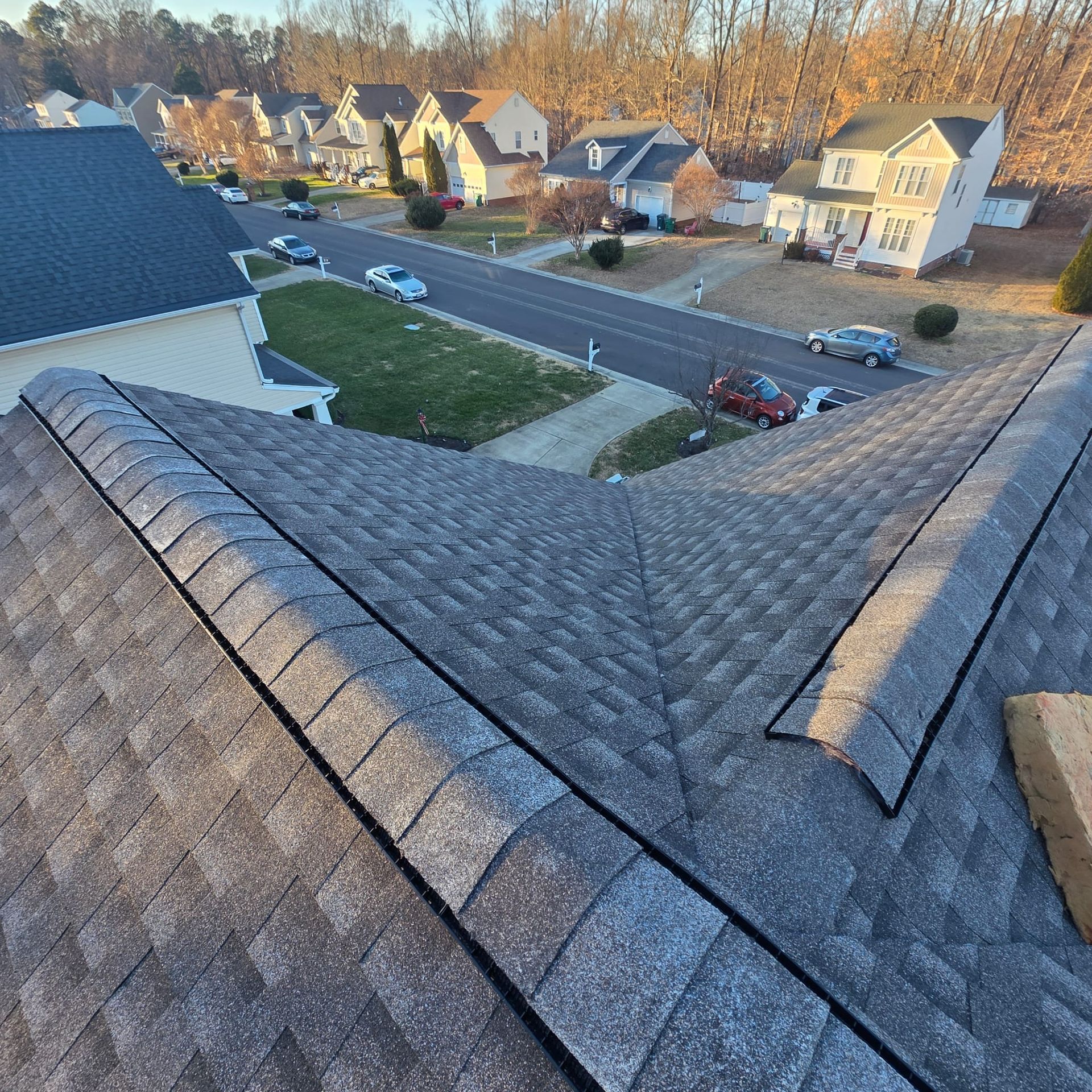 Overhead view of a house roof, asphalt shingles in shades of gray, with a neighborhood street and houses in the background.