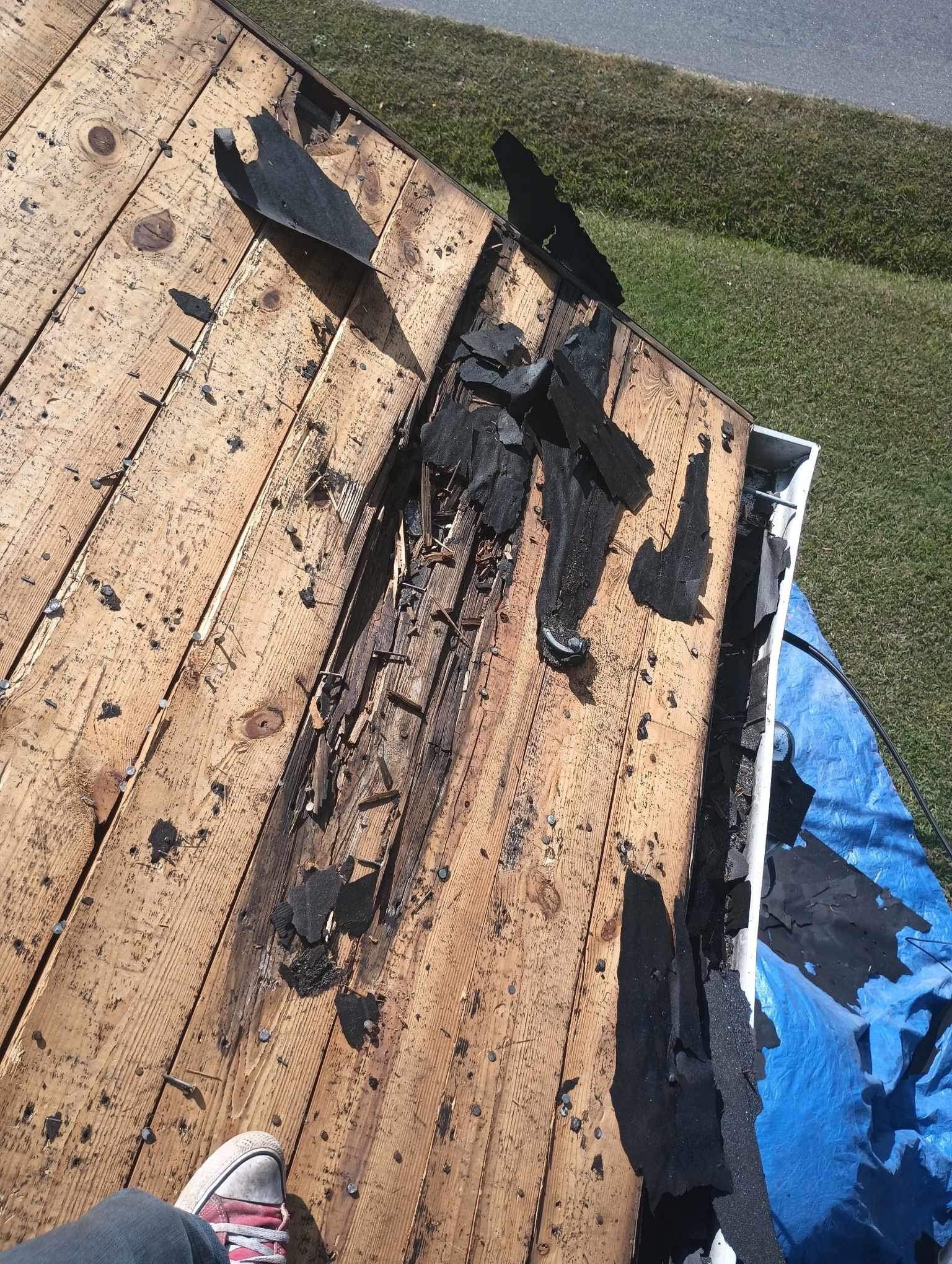 Damaged roof with torn black shingles and exposed wood; a person's foot in a sneaker is visible.