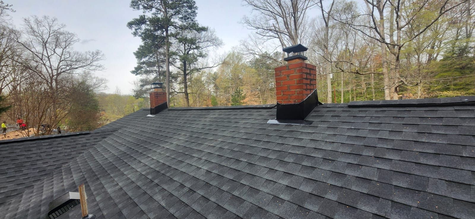 Two brick chimneys on a shingled roof with trees in the background.