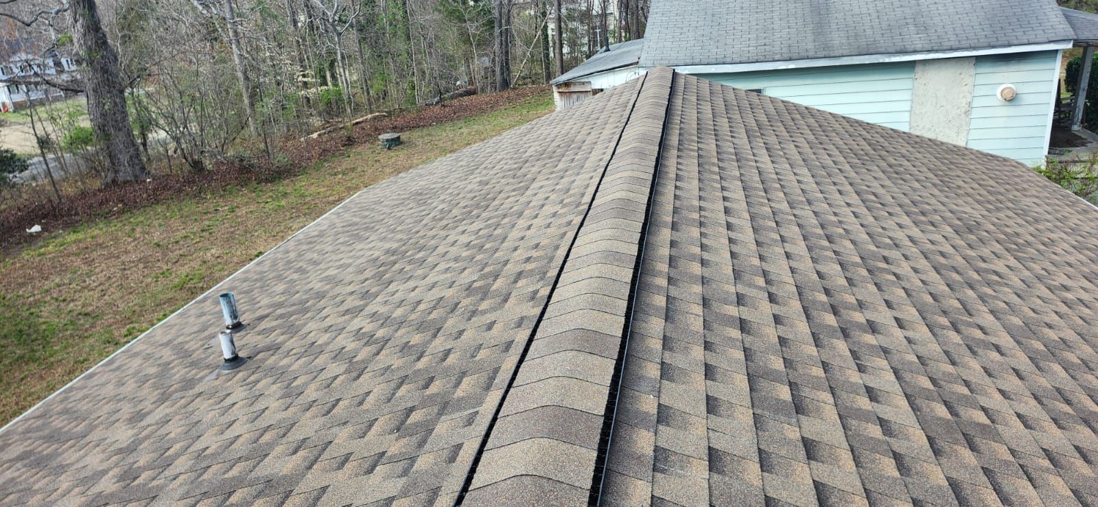 View of a brown shingled roof with a central ridge, trees and grass in the background.