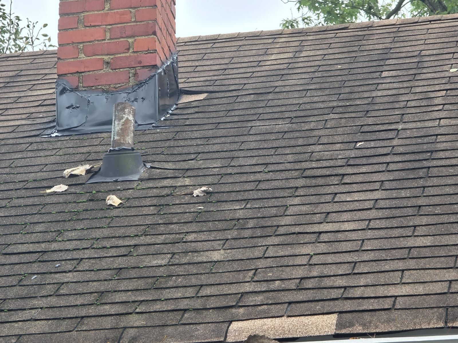 Brown shingled roof with a brick chimney. Flashing around the chimney shows signs of wear and tear, and some shingles are missing.