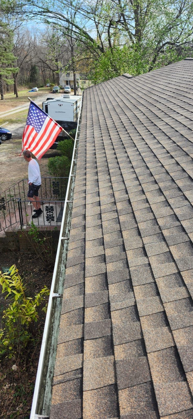 Person holding American flag by a house with a shingled roof and gutters, sunny day.
