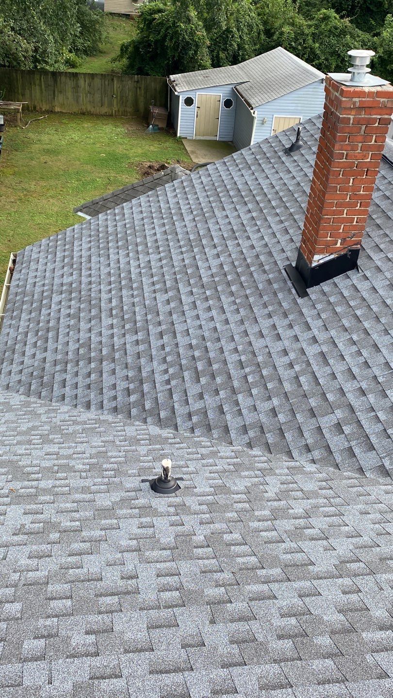 Gray shingled roof with a brick chimney and small shed in the background. Green grass and trees visible.