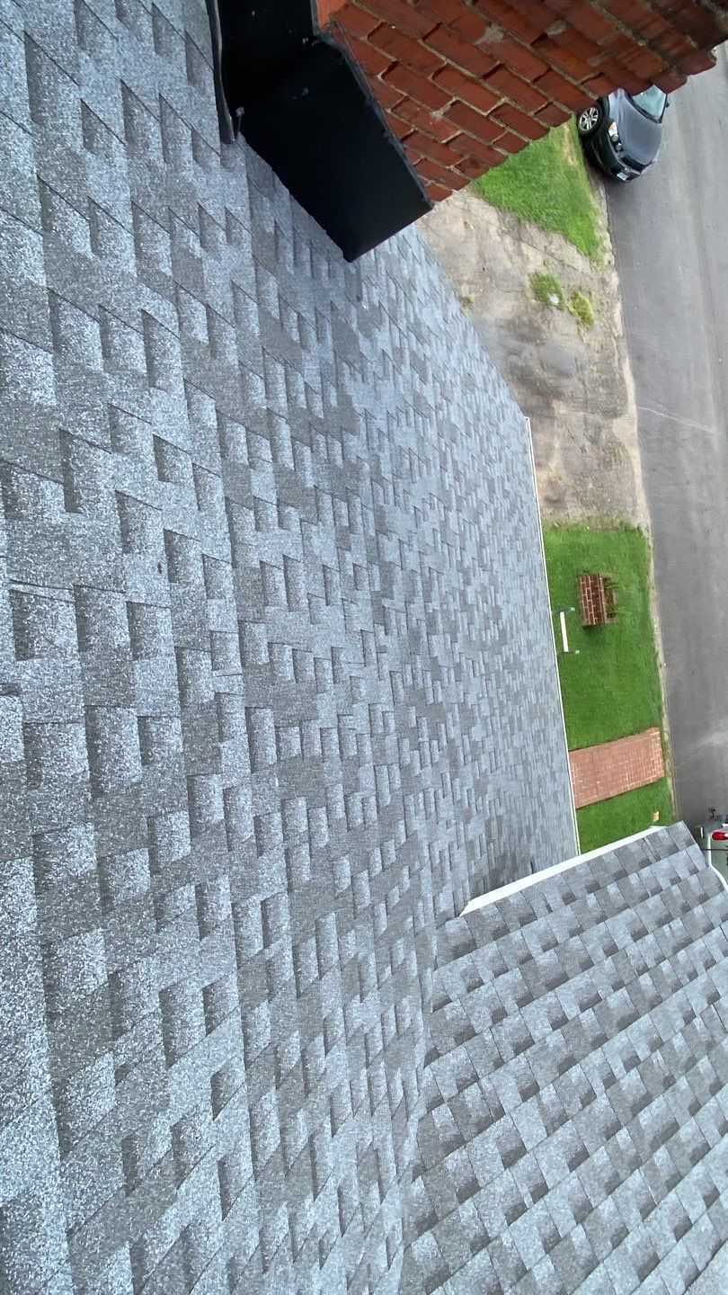 Gray asphalt shingle roof on a house, angled view from above. Lawn and brick trim visible.