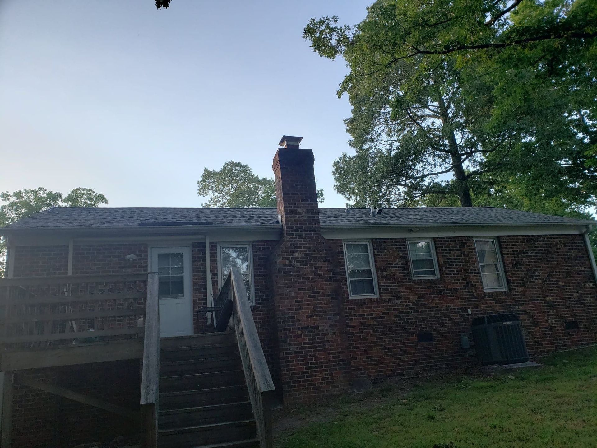 Brick house exterior with a chimney, a deck, and stairs, set in a yard with trees.