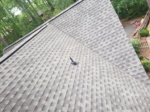 Gray asphalt shingle roof on a house, with a vent and trees in the background.