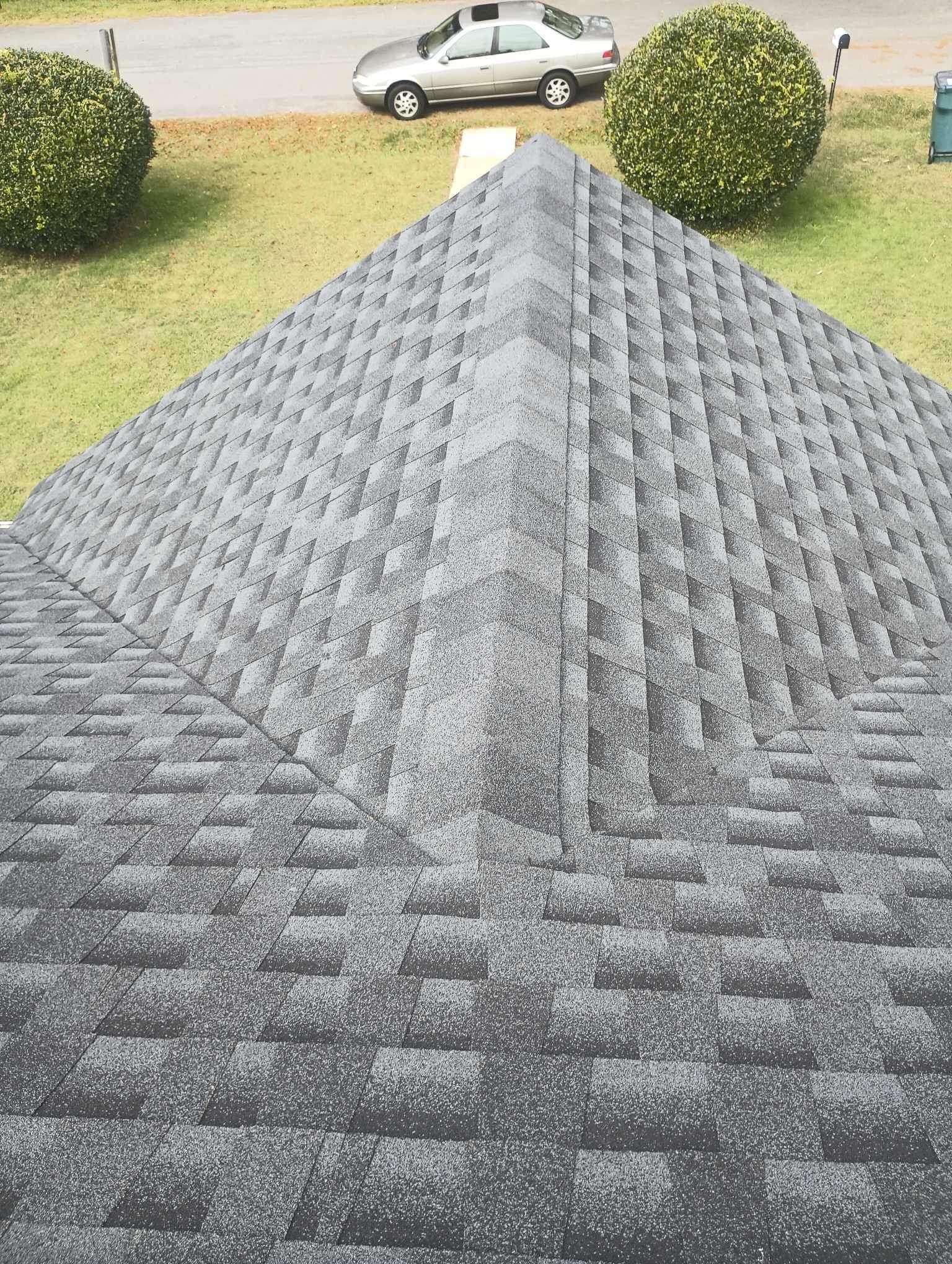 View from roof apex. Gray asphalt shingle roof with a central ridge. Green lawn, car, and bushes in the background.