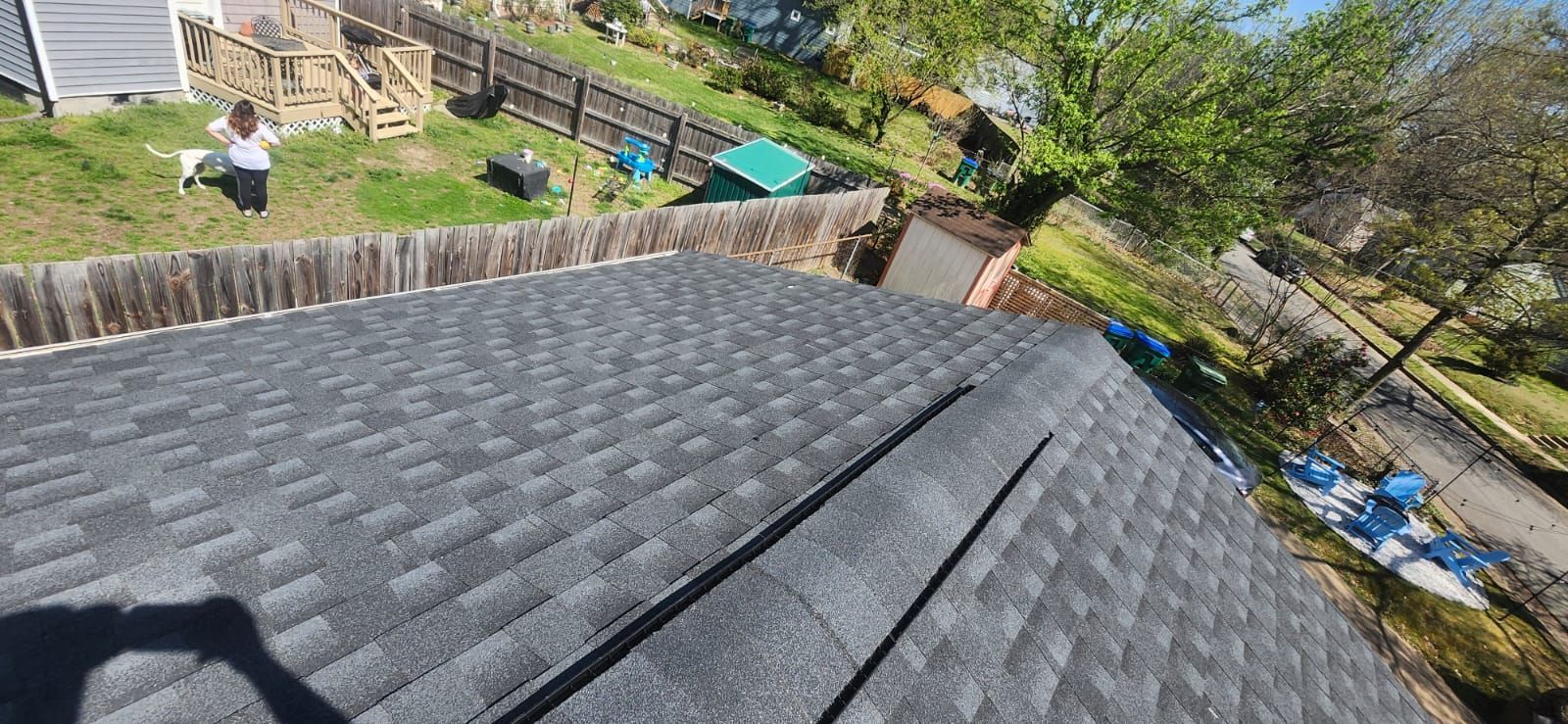 A view of a house roof with new asphalt shingles, overlooking a backyard with trees and a fence.