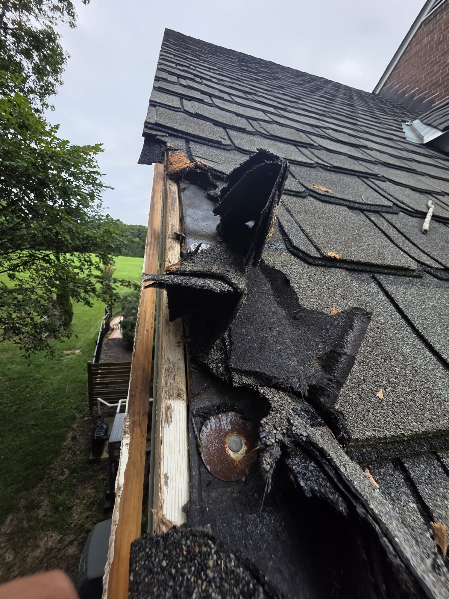 Damaged roof corner with missing and torn shingles, showing wood and metal flashing against a cloudy sky.