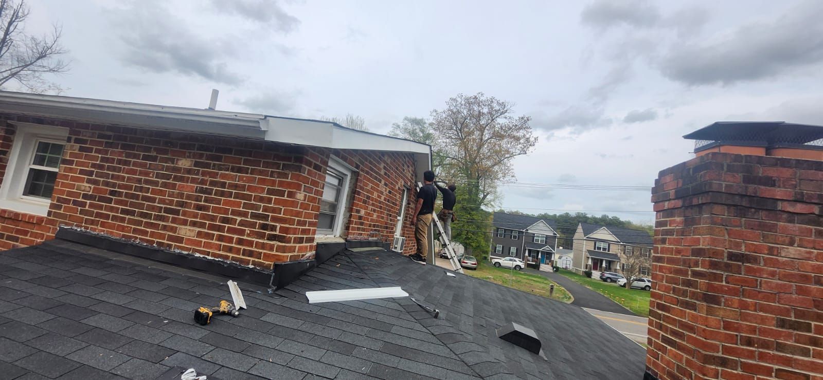 Roofers on a brick building with a dark shingled roof, cloudy sky.