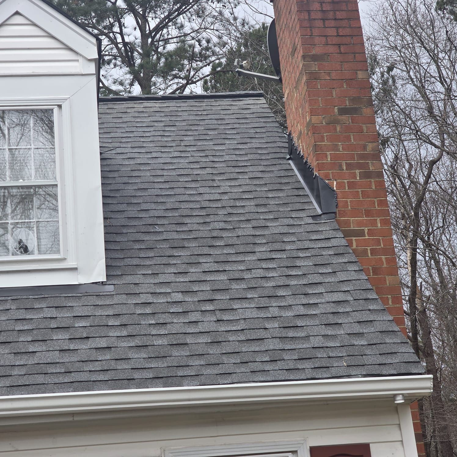 Gray asphalt shingle roof of a house with a brick chimney and white trim.