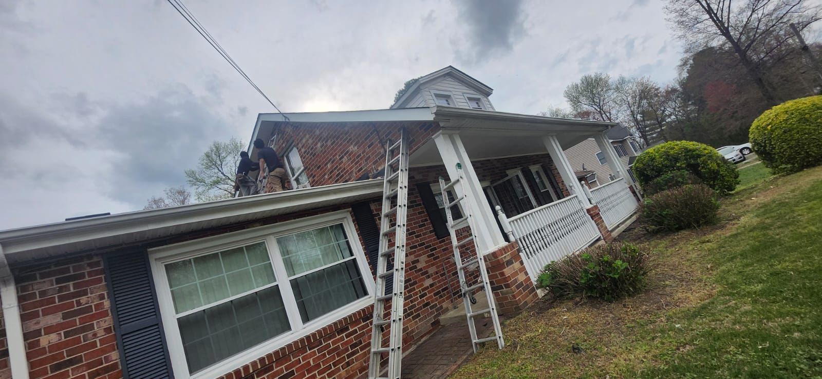 House with red brick and white trim, two ladders leaning on it. Overcast sky, front yard on a slope.