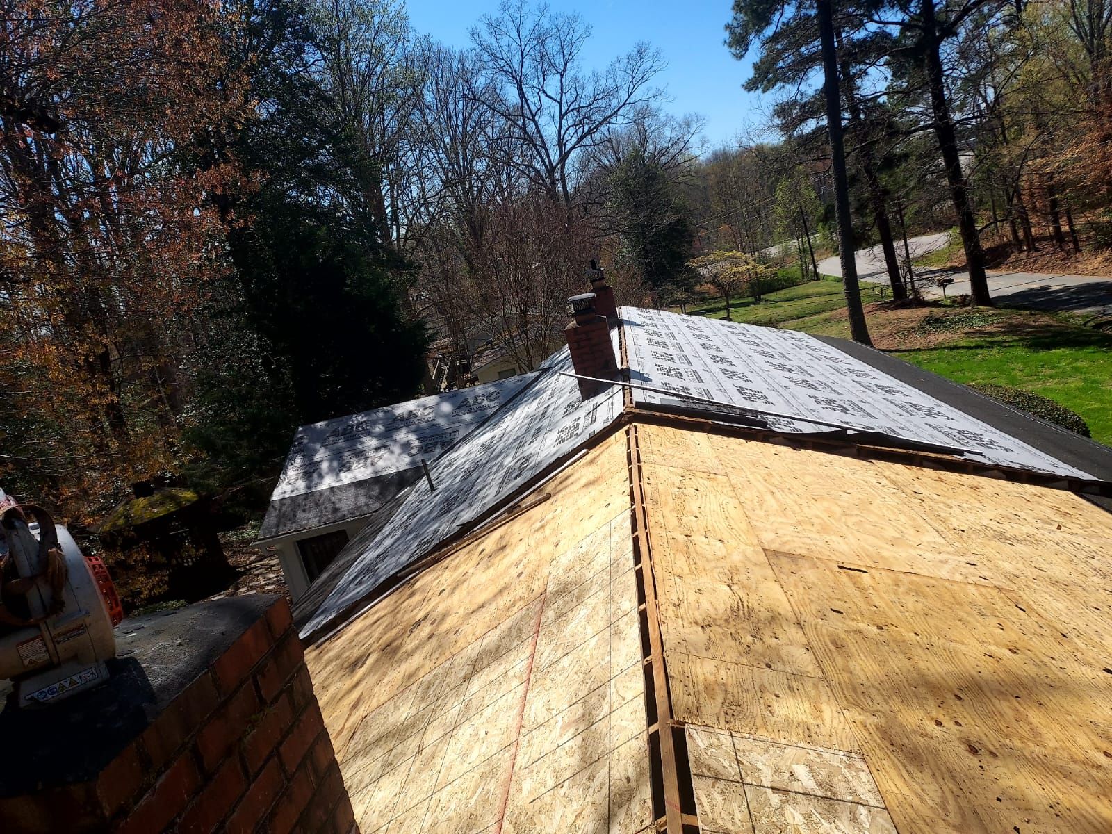 Roof partially covered with old shingles and exposed plywood, with a brick chimney and treeline.
