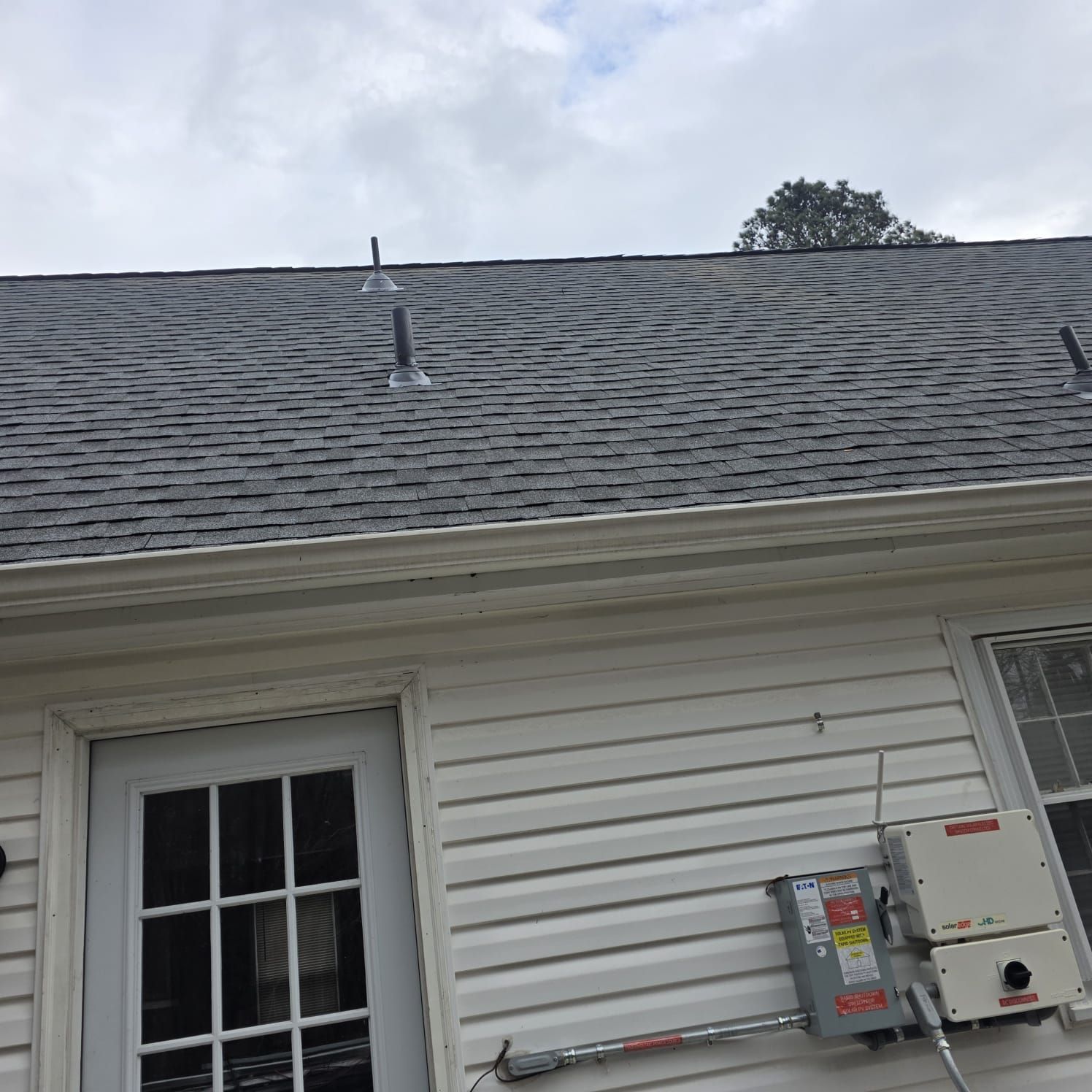 Gray shingled roof with vent pipes, white siding, and electrical equipment mounted on the exterior of a building.