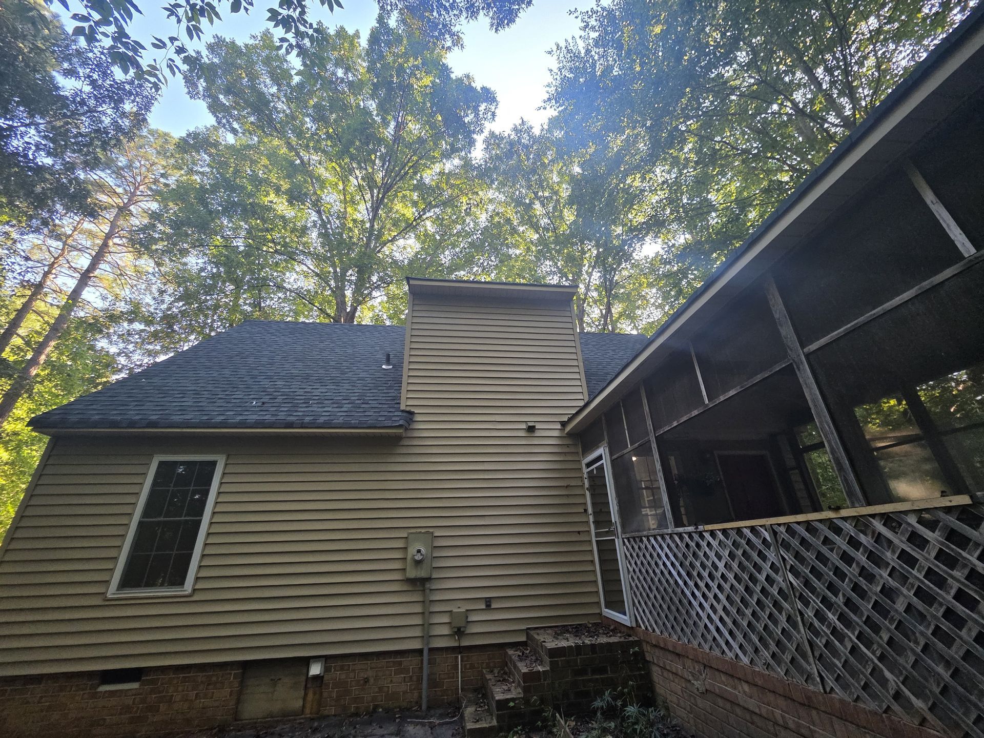 Tan house with a chimney and screened porch, surrounded by trees under a blue sky.
