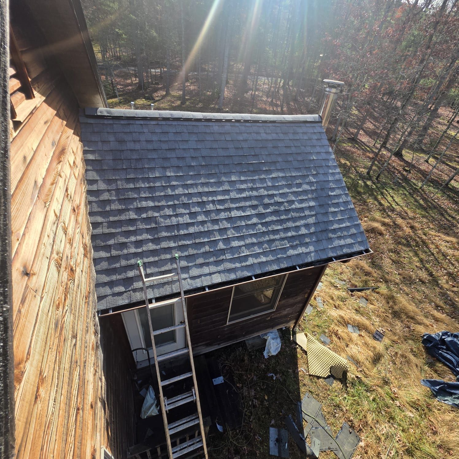 A house with dark shingles, a chimney, and a ladder against a small roof extension; sunny outdoor setting.