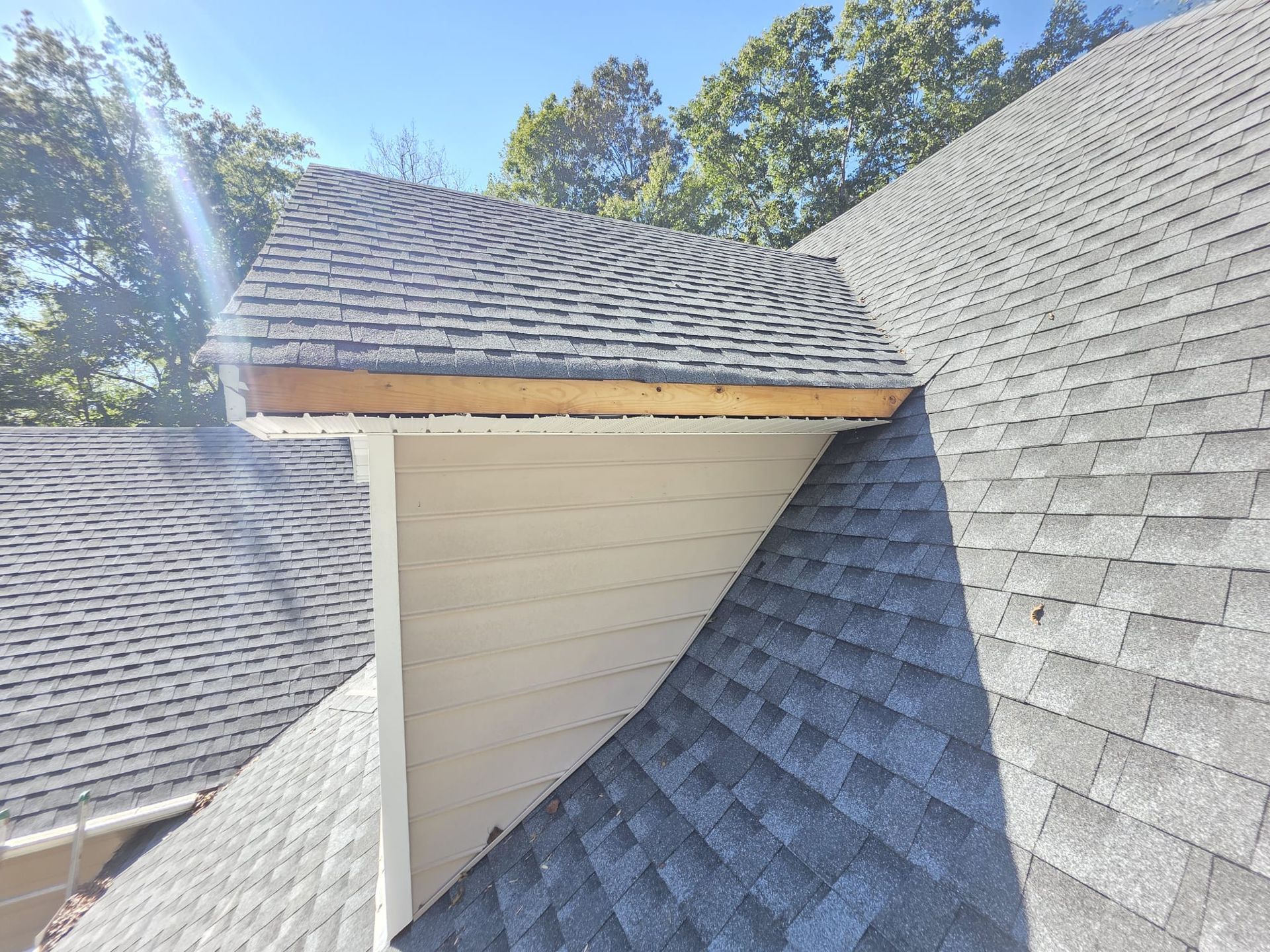 Close-up of a roof corner showing gray shingles, exposed wooden trim, and white siding, with a clear blue sky.
