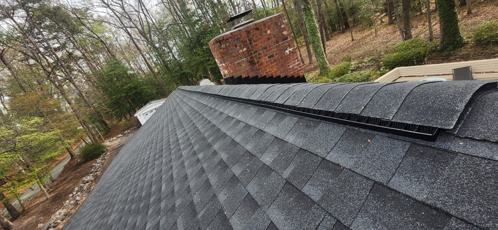 Roof with dark gray shingles, leading up to a brick chimney. Trees in the background.