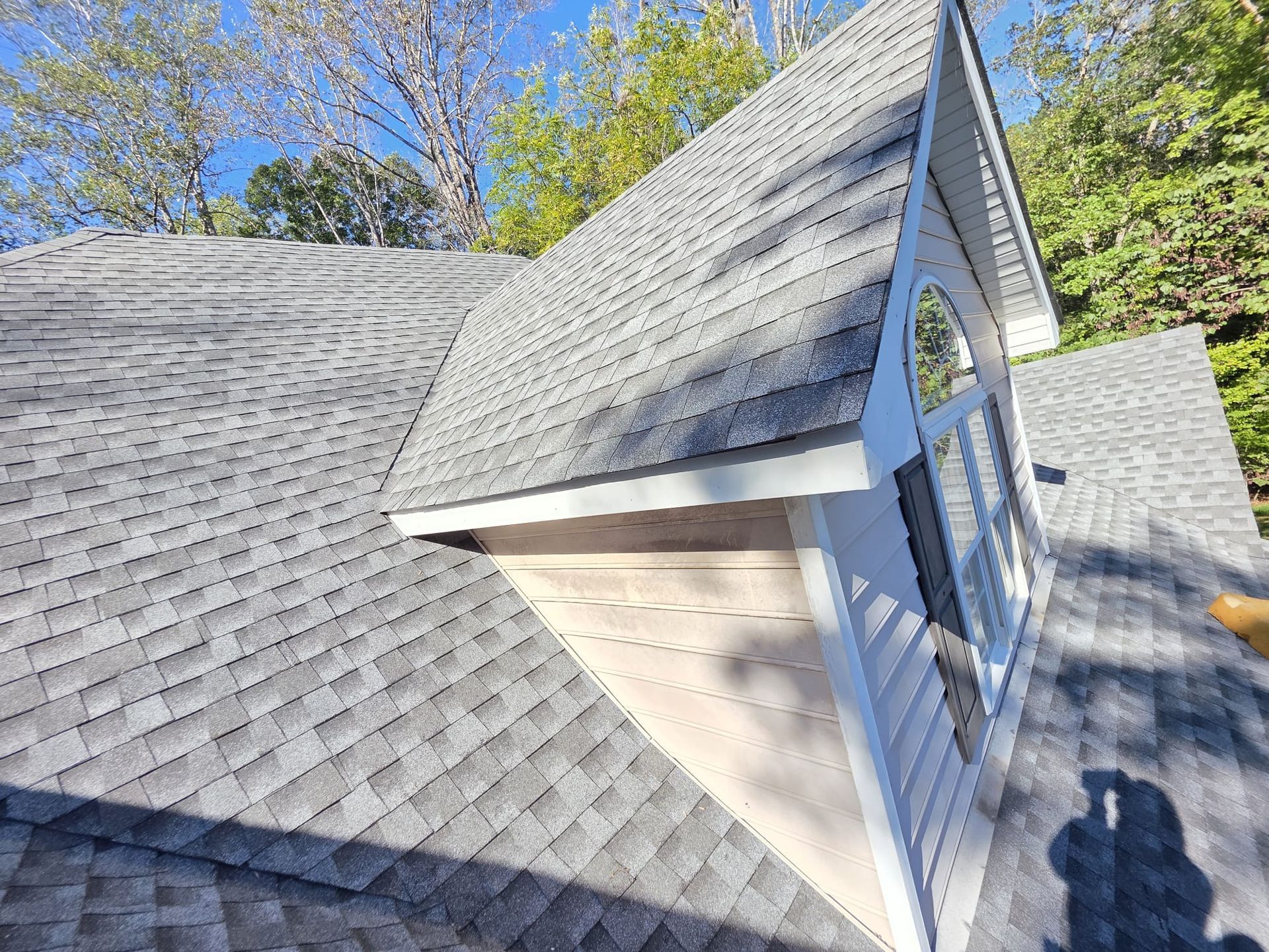 Gray shingled roof with a white gable window, viewed from above, surrounded by trees.