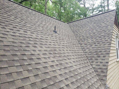 Brown shingle roof of a house with a vent pipe, set against green trees.