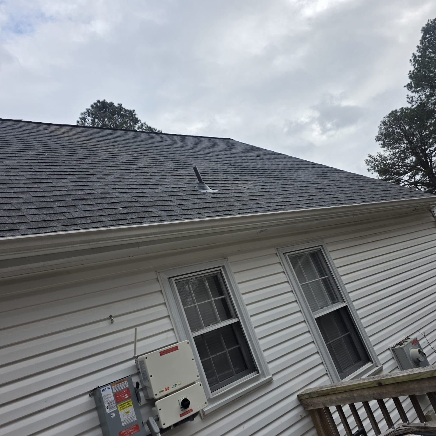 Gray shingled roof on a white house with two windows and a cloudy sky.
