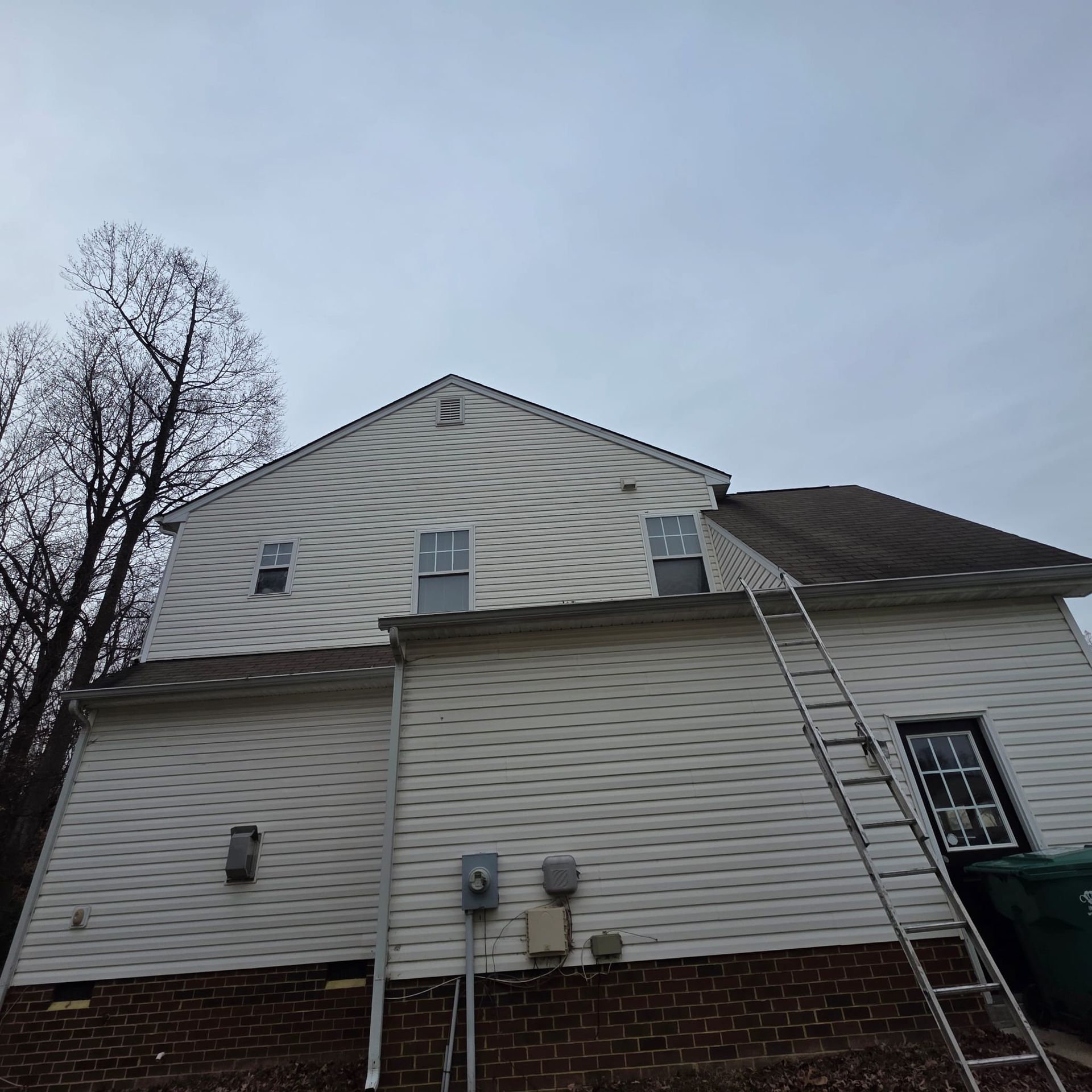 Two-story house with white siding and a ladder leaning against the roof, set against an overcast sky.