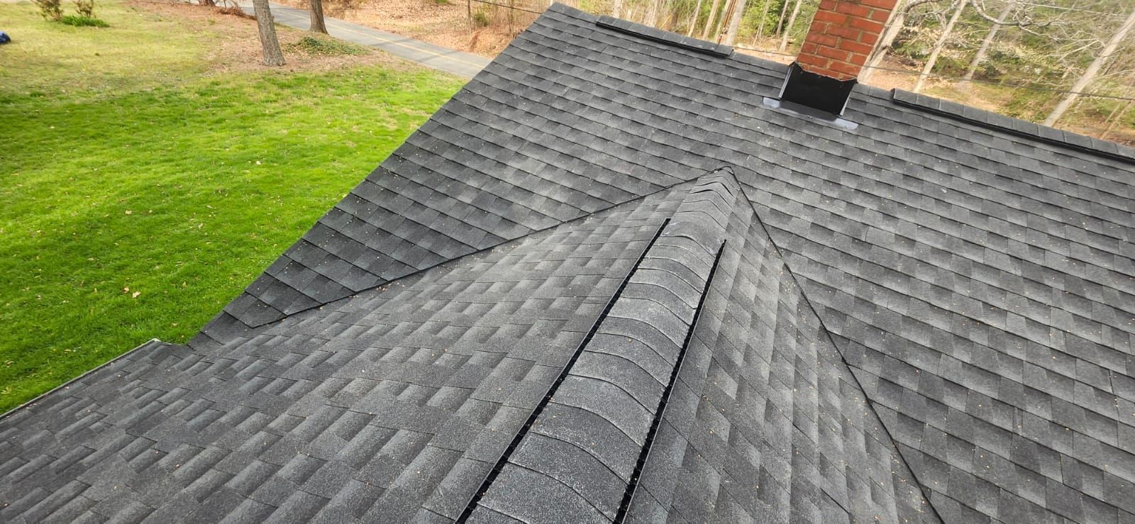 Overhead view of a dark gray shingled roof with a chimney, green grass, and trees in the background.