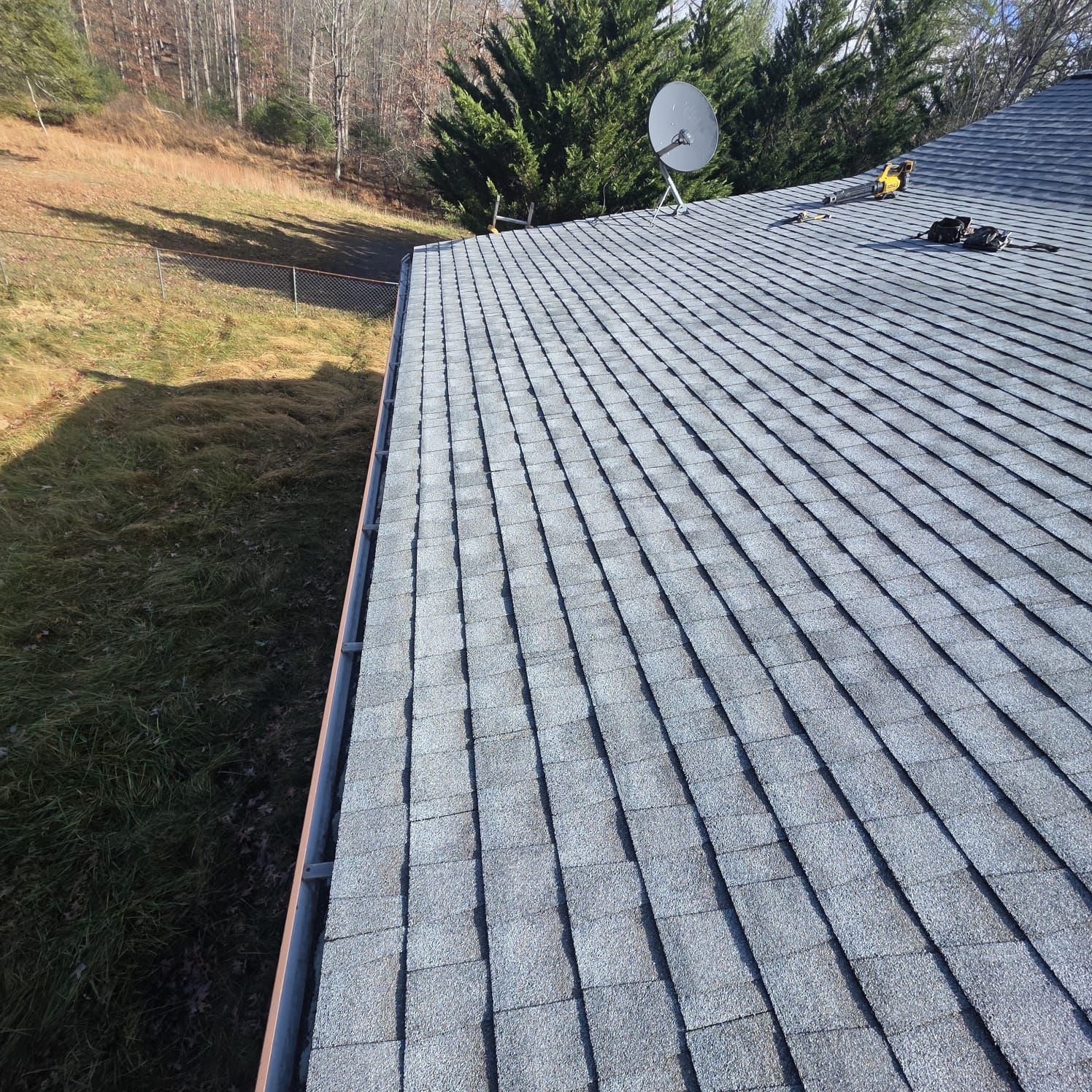 Gray shingle roof with a satellite dish, against a grassy hill and trees.