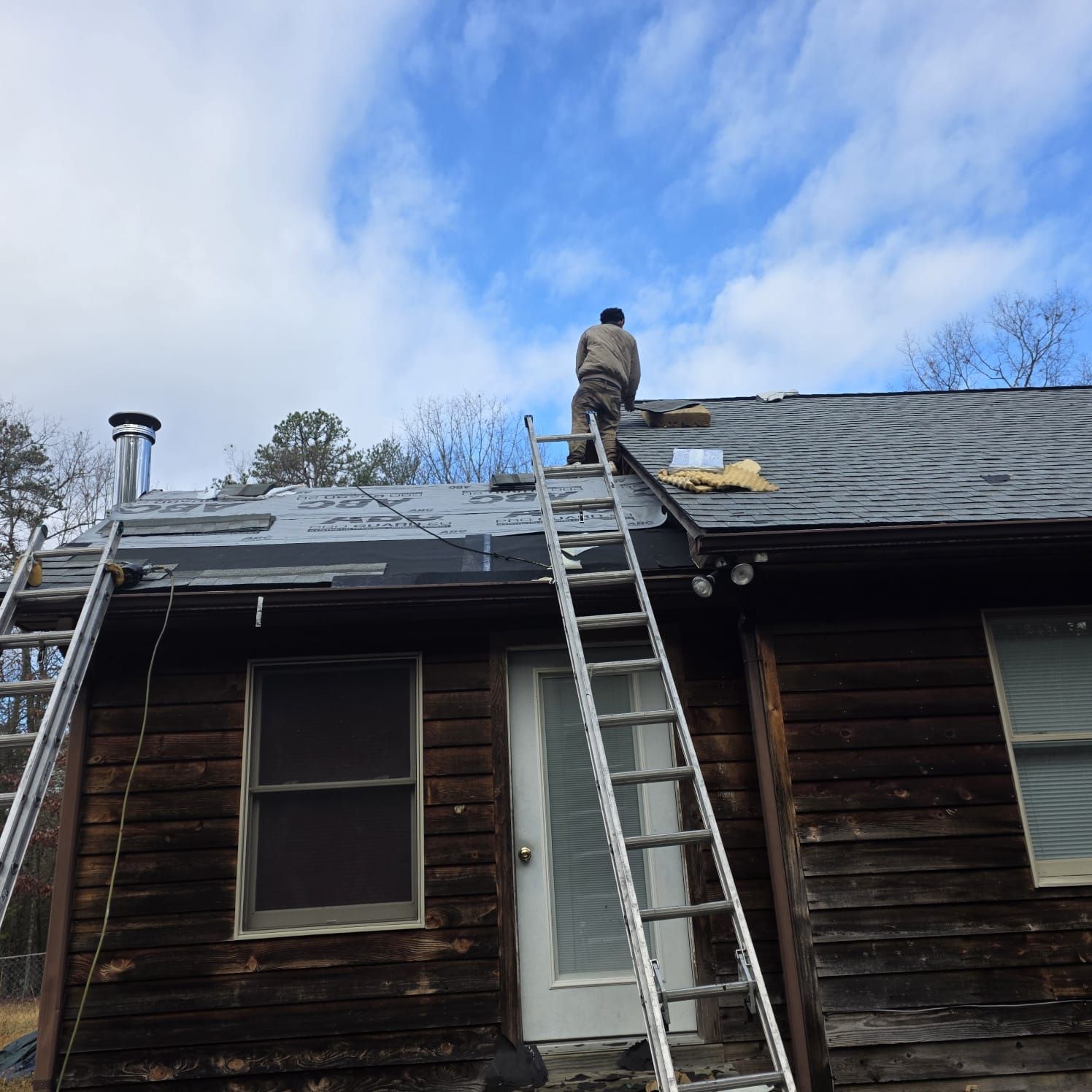 Person on a ladder, working on a dark roof. Cloudy blue sky. Wooden house.