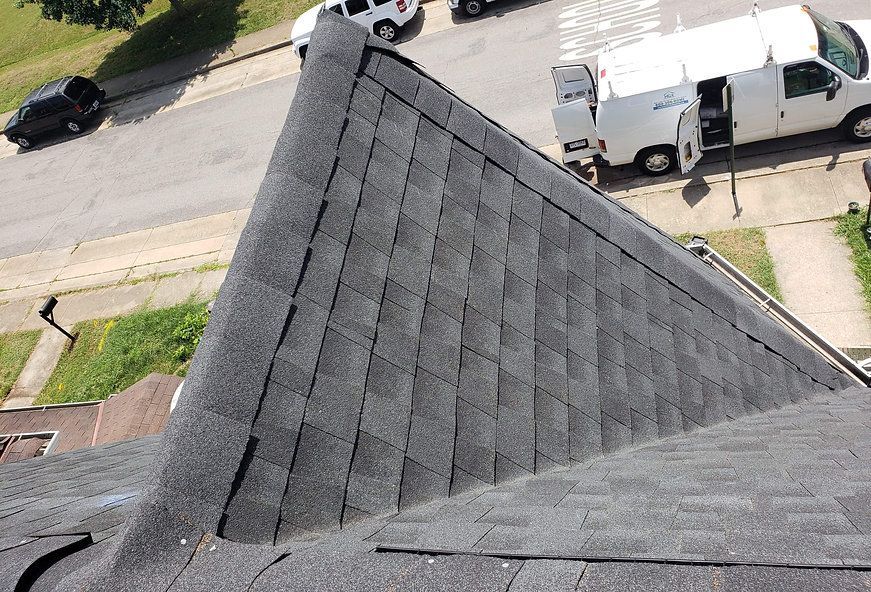 Dark asphalt shingle roof on a house, angled view, street and parked vehicles in the background.