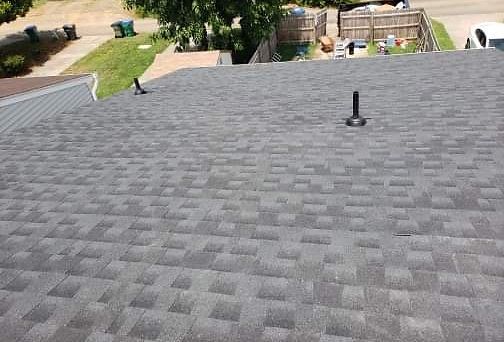 Gray shingled roof with a black pipe, overlooking a grassy yard and houses.