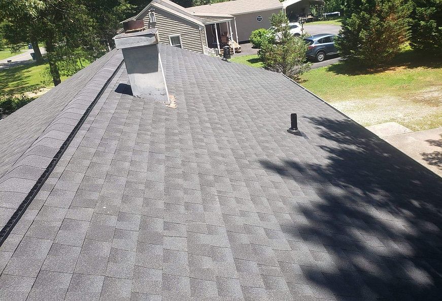 Gray asphalt shingle roof on a house, with a chimney and vent pipe. Trees and grass in the background.
