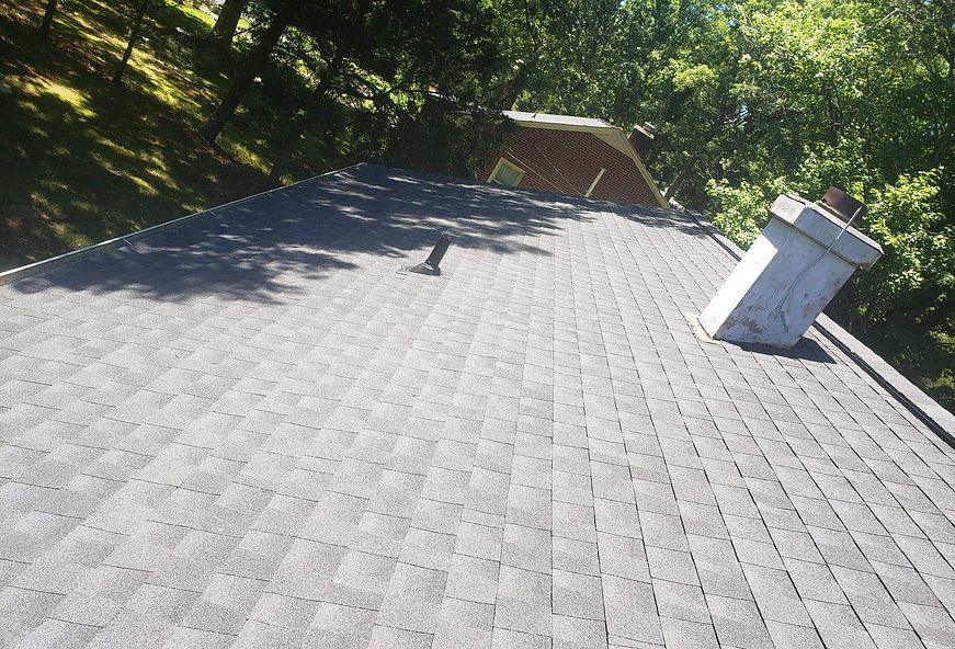 Gray shingled roof with a chimney and part of another roof visible. Trees in the background.