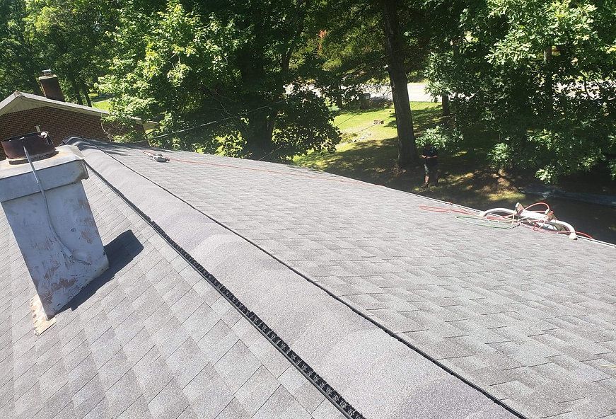 Gray shingled rooftop with a chimney, a long vent, and trees in the background on a sunny day.