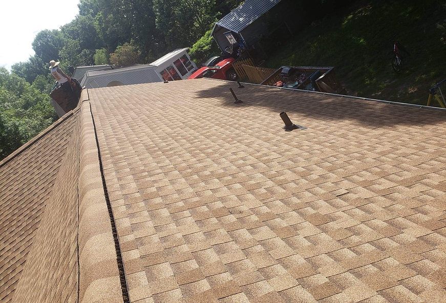 Brown shingled roof with a dark edge, trees, and partial view of two vehicles on a hillside.
