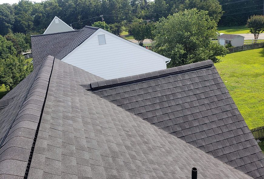 A house roof with dark gray shingles, viewed from above, surrounded by green trees and grass.