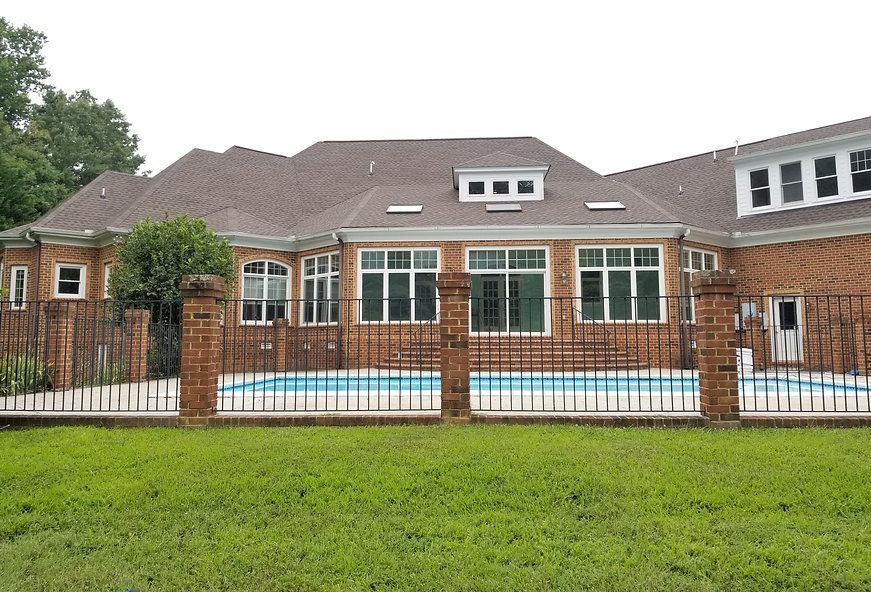 Brick house with a pool, enclosed by a wrought iron fence. Green grass in the foreground.