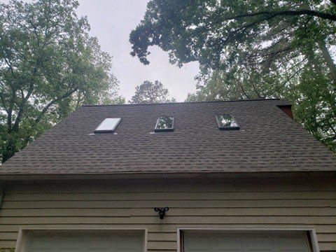 Garage roof with three skylights, covered in asphalt shingles, under cloudy sky.