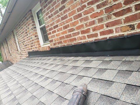 Roof with asphalt shingles next to a brick wall. A black flashing sits between.