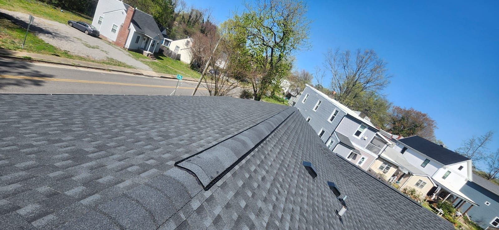 View of a dark gray asphalt shingle roof, with a black vent in the center, and houses and trees in the background.