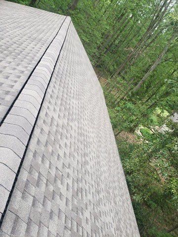 Gray asphalt shingle roof, with a black ridge line, against a backdrop of green trees.