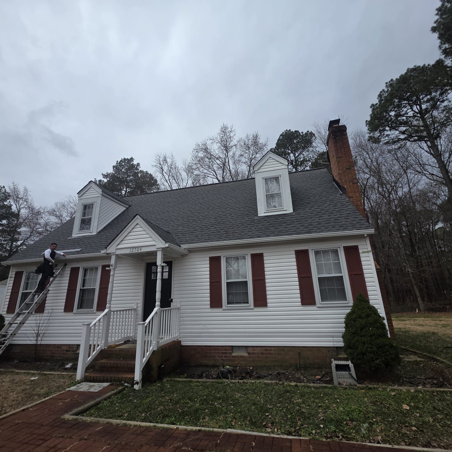 White house with dark gray roof, red shutters, two dormers, and brick chimney. A person on a ladder. Cloudy sky.