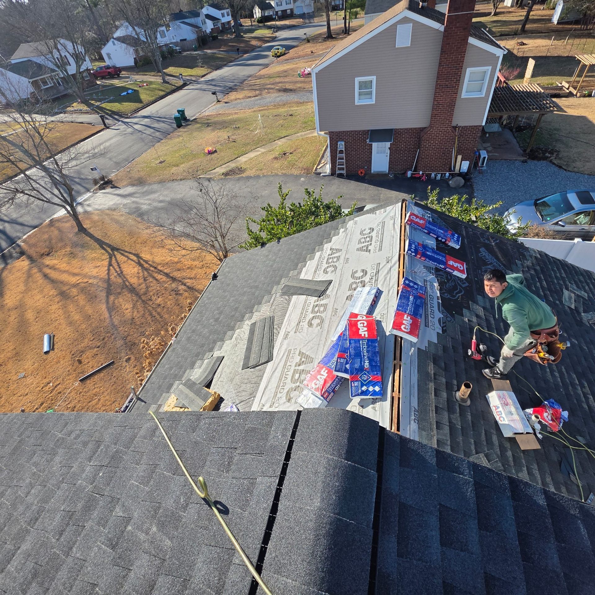 Roofer installing shingles on a house. The house is brown, and the surrounding area has houses and trees.