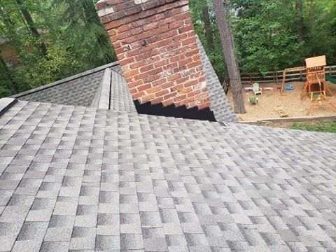 Gray shingle roof with a brick chimney in a backyard setting with trees and a playset.