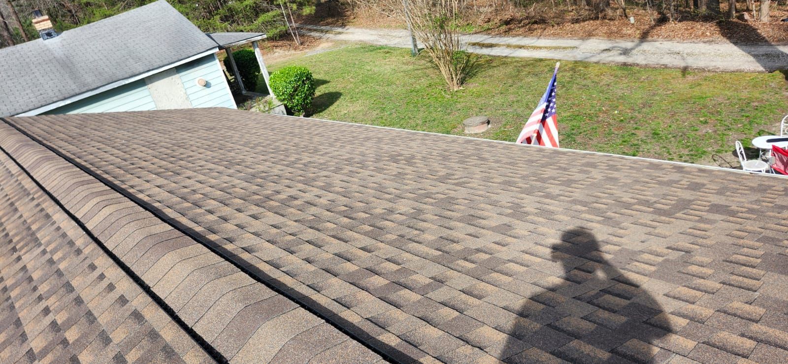 Brown shingled roof with a shadow, the American flag, a green yard, and a house are in view.