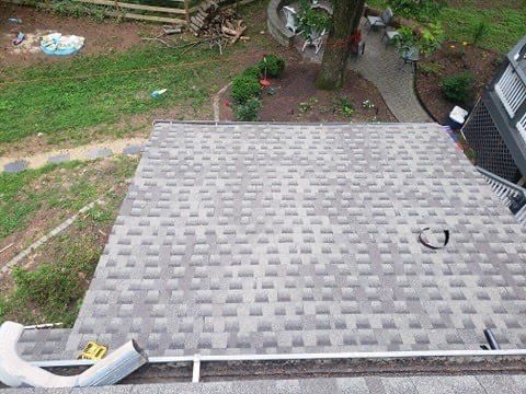 View of a residential roof covered in grey shingles, gutter and downspout at the edge, surrounded by greenery.