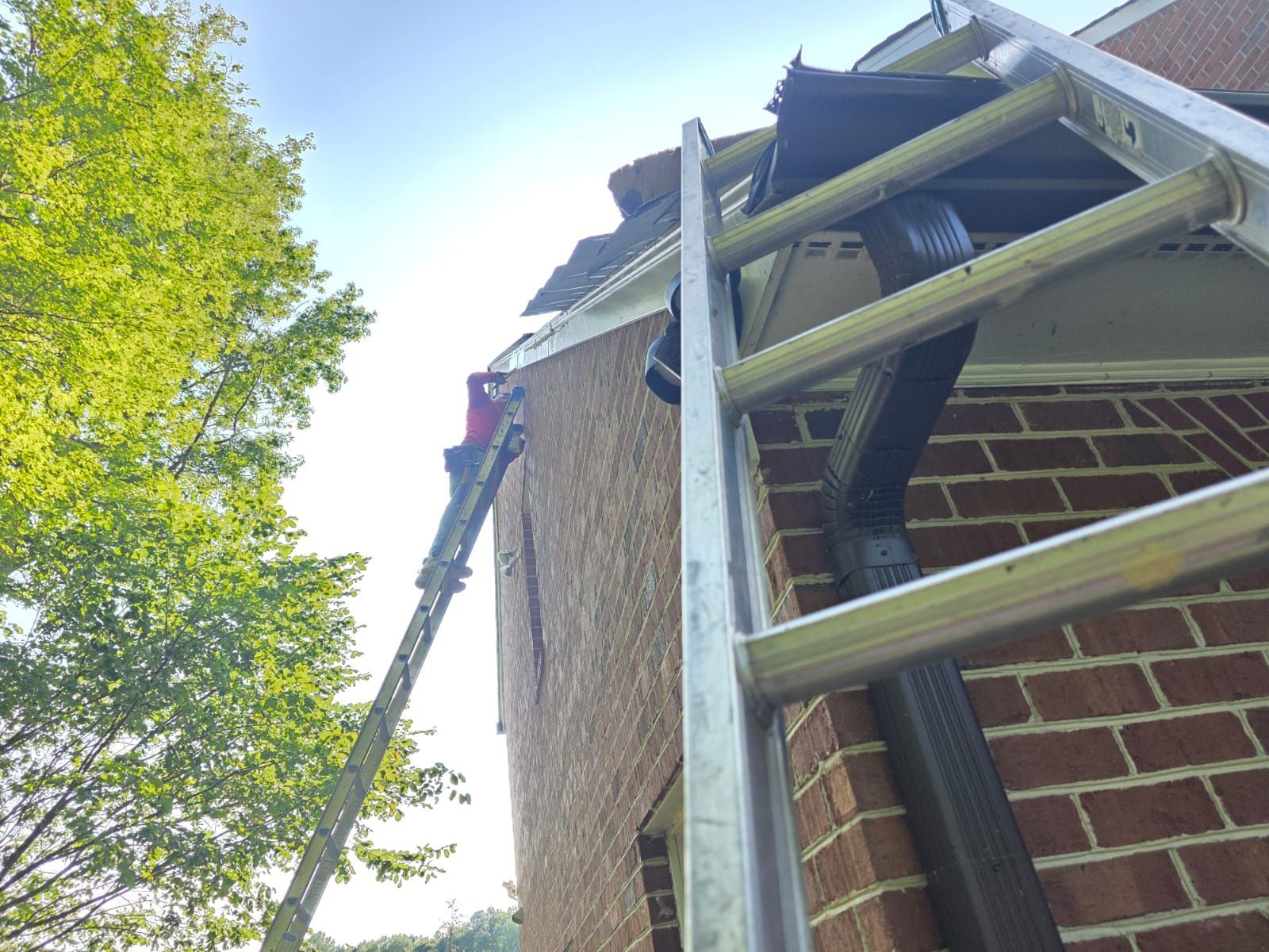 Ladder leaning against brick building; person on roof, debris visible. Blue sky, green trees.